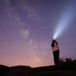 Una mujer observando las estrellas en la noche usando una linterna en la cabeza parada en el tope de una montaña.