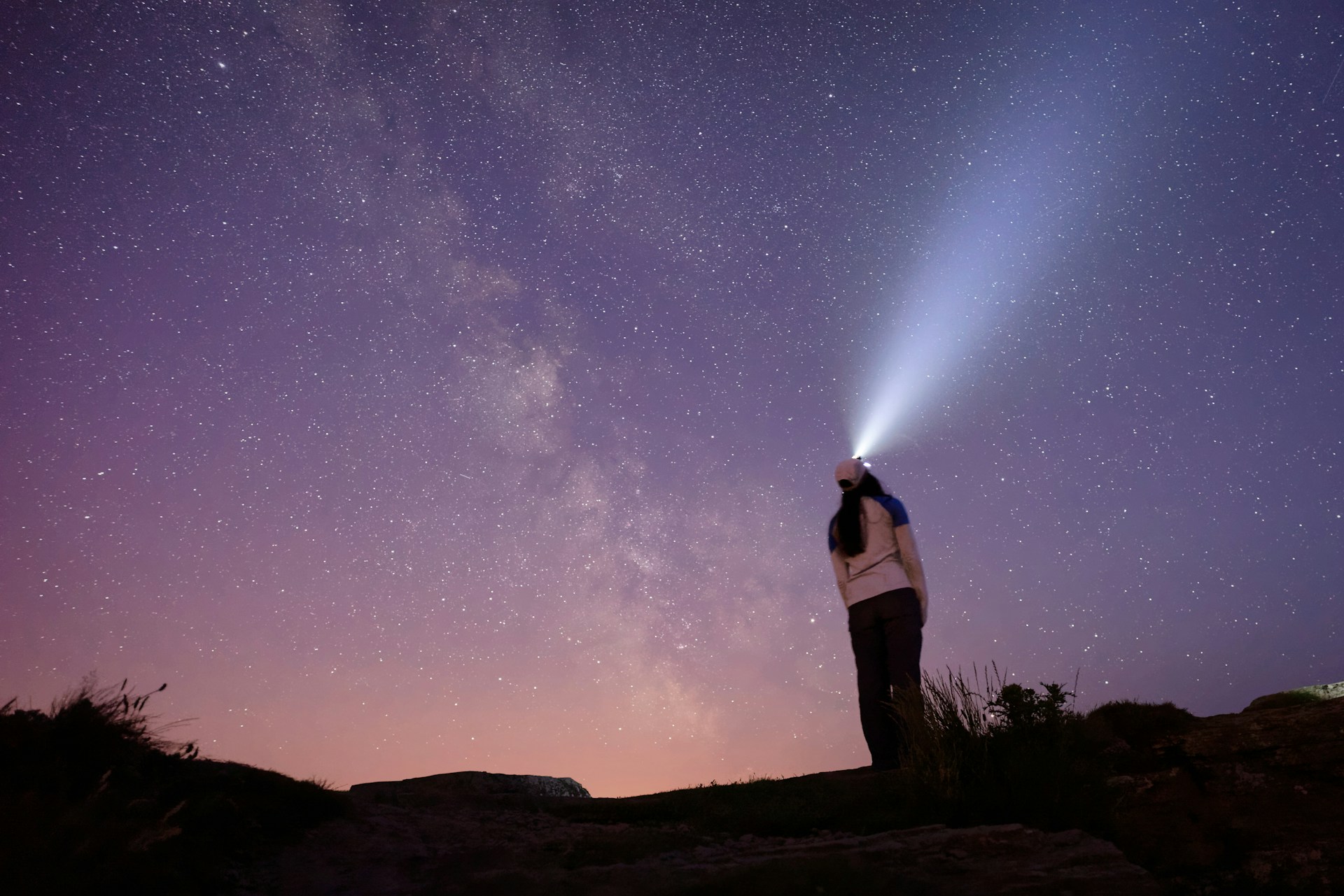 Una mujer observando las estrellas en la noche usando una linterna en la cabeza parada en el tope de una montaña.