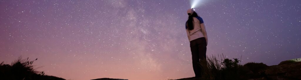 Una mujer observando las estrellas en la noche usando una linterna en la cabeza parada en el tope de una montaña.