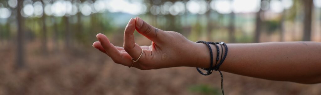 Una mano con pulseras en un bosque verde desolado con un piso en tierra haciendo una pose que se refiere a pensar.