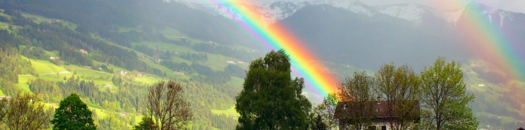 Arcoiris doble formándose detrás de unas casas y frente a unas montañas con nieve.